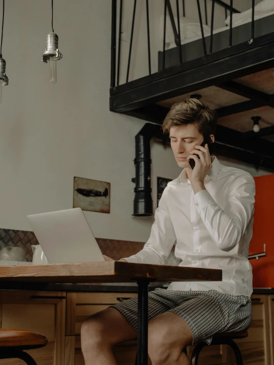 young man talking on his cell phone at home, while sitting at a table using laptop