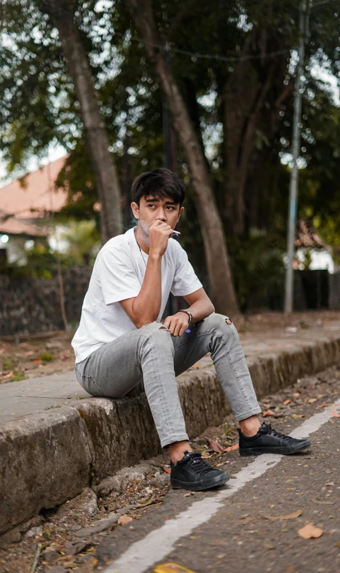 a young man sitting on the curb and leaning against his knee, as if talking on a cell phone