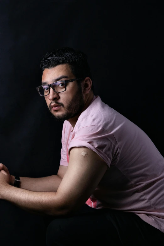 a man in a pink shirt and glasses sitting against a black background