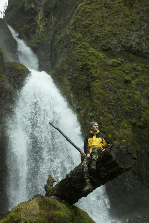 a man in yellow shirt sitting on a tree nch by a waterfall