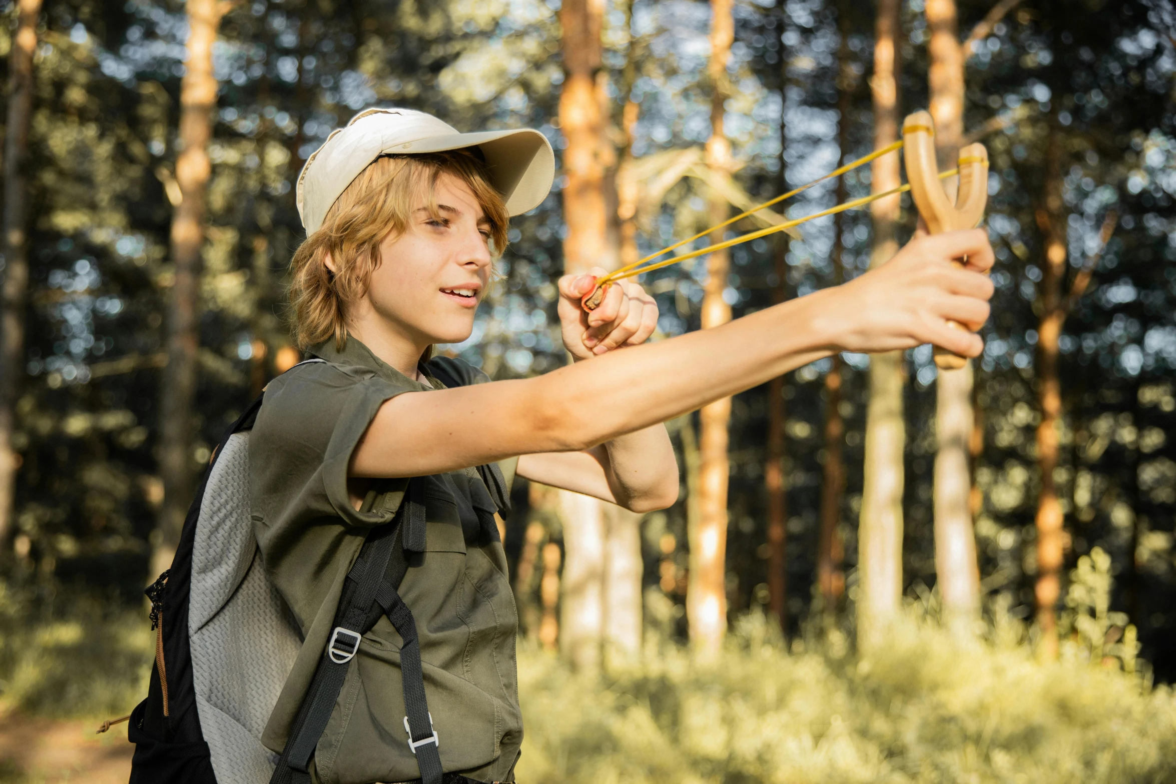 a boy in a hat and suspenders is holding a stick in a forest