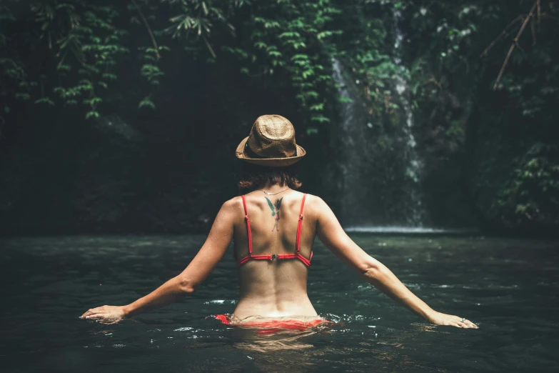 woman with back turned, with hand painted on, sitting in water in front of waterfall