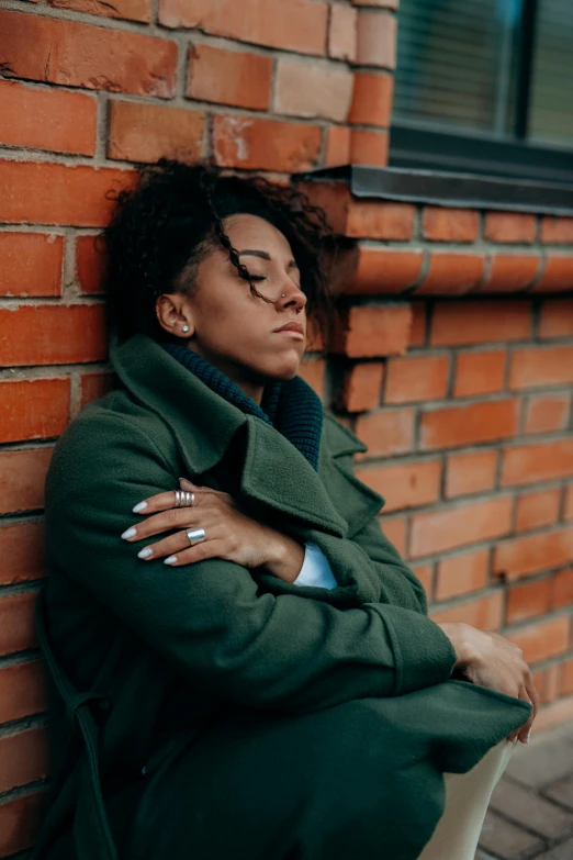 a woman leaning against a brick building wearing a coat and sitting on the side of the street