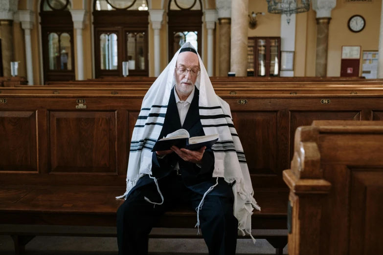 a man in black outfit sitting on pew with a white and black stole over his head