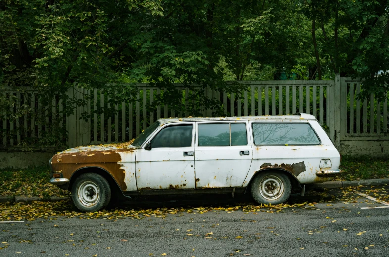 a rusted, car sits in a parking lot surrounded by trees