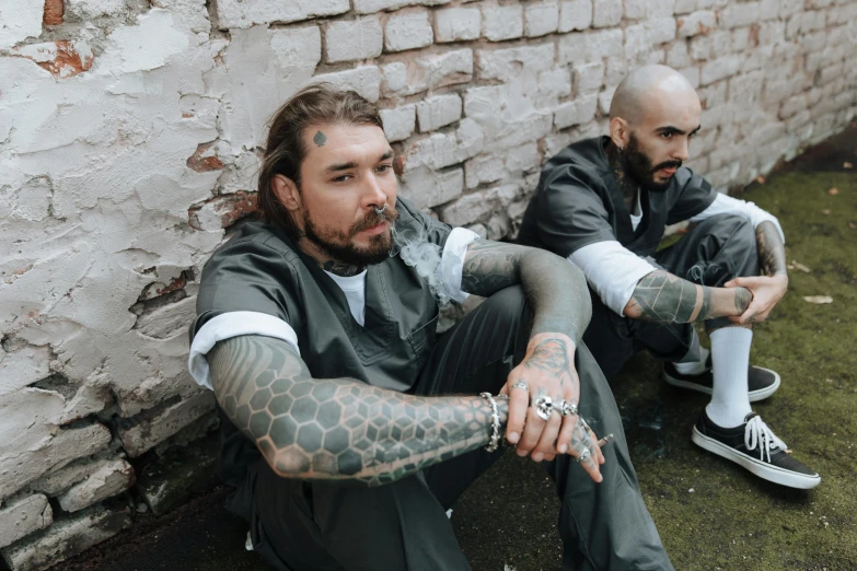 two men wearing tattoos are sitting against a brick wall