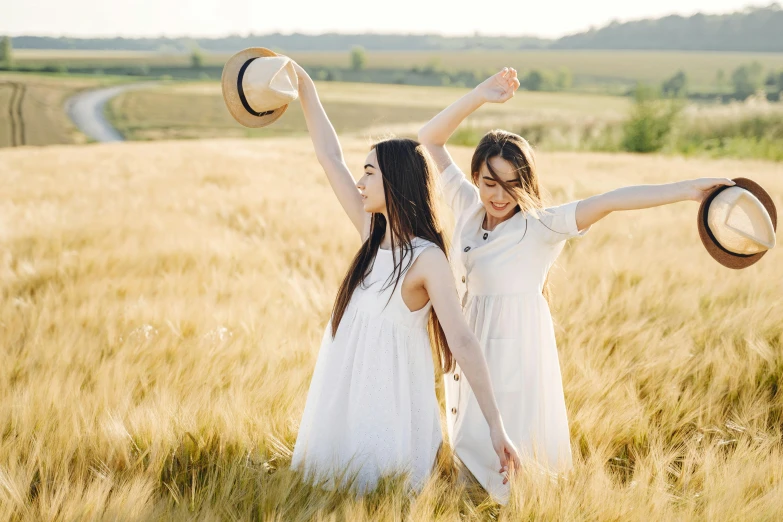 two girls stand together in a field holding hats