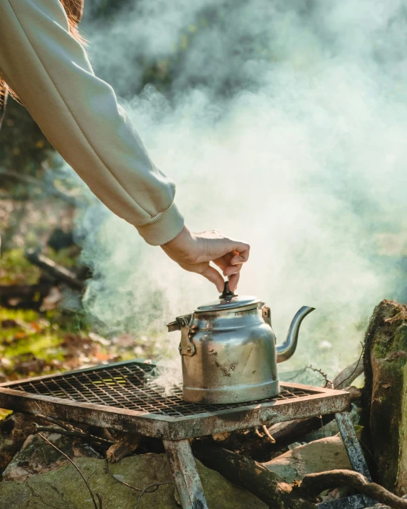 the person holds a kettle over a camp stove while cooking