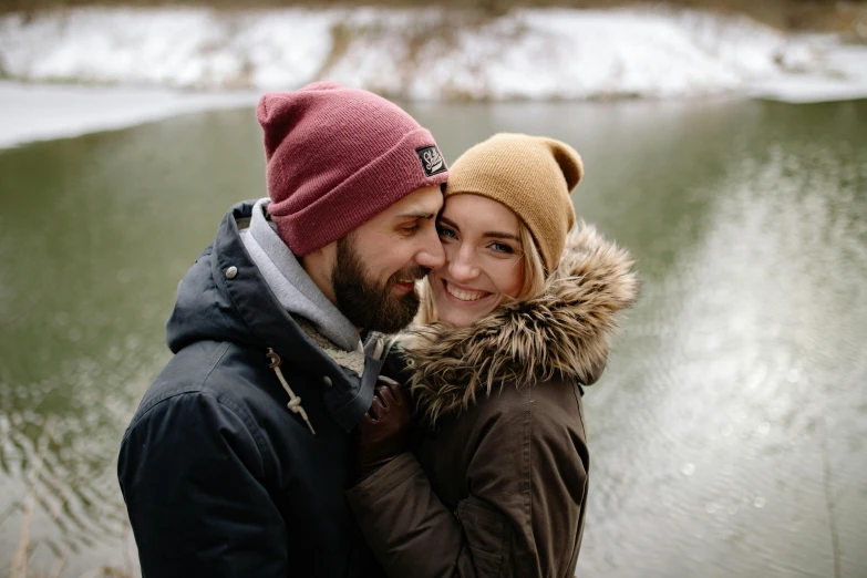 a man and woman in winter attire stand by a body of water with a mountain view in the background