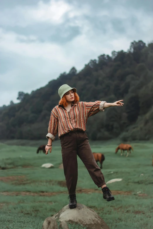 a woman with a hat, red hair and brown pants standing on a rock in a field