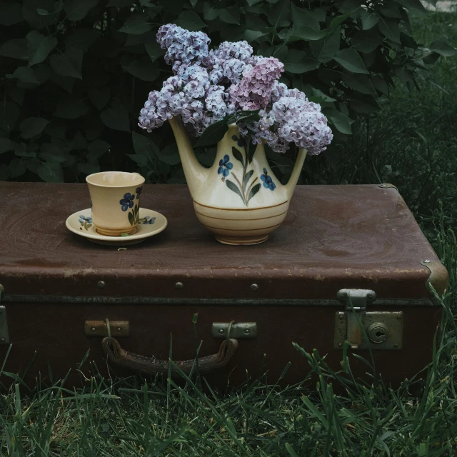 purple flowers in a vase in front of a teacup on an old suitcase