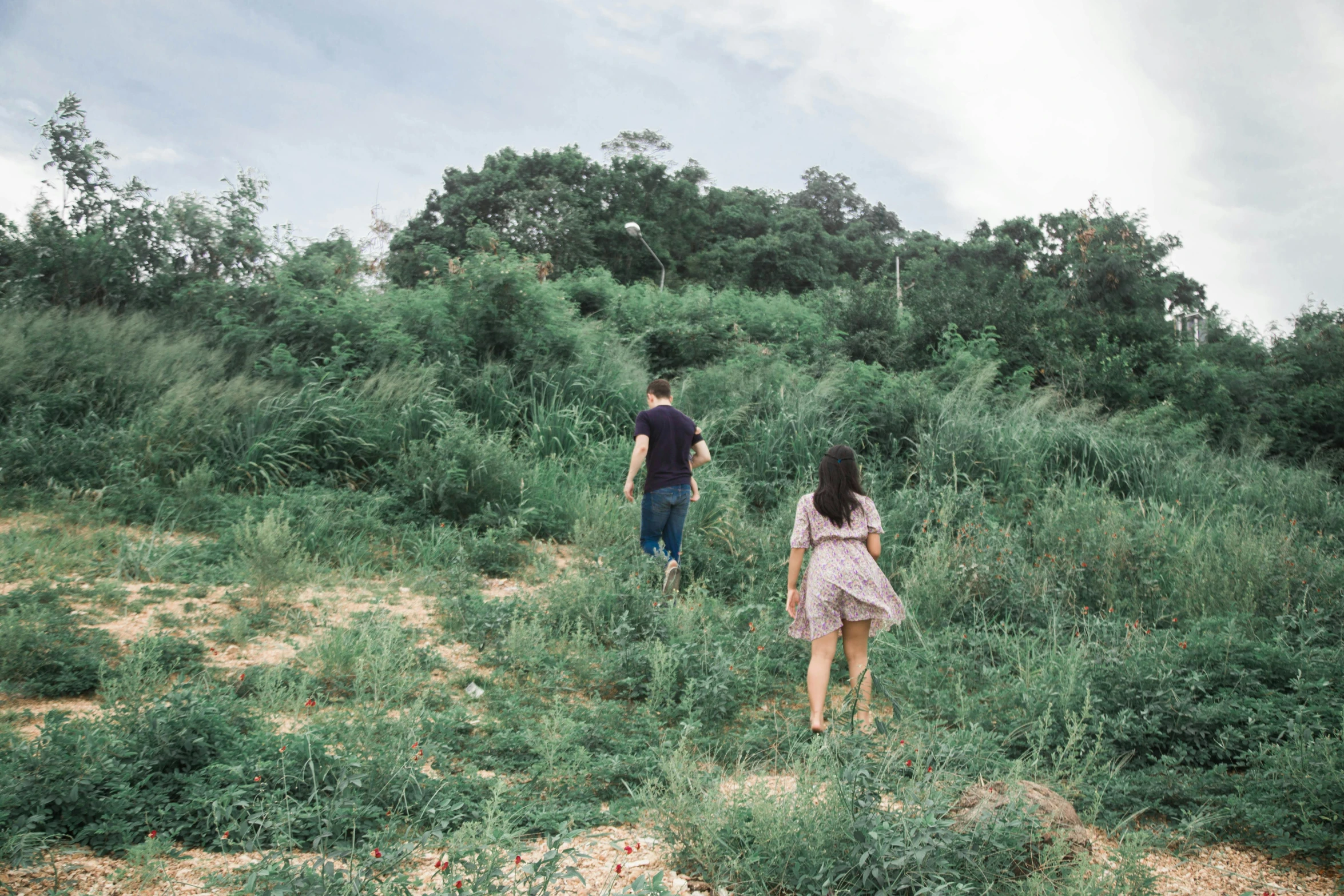 two people walk through the field next to green brush