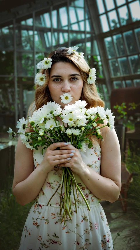 a woman in a dress holding flowers with a greenhouse in the background