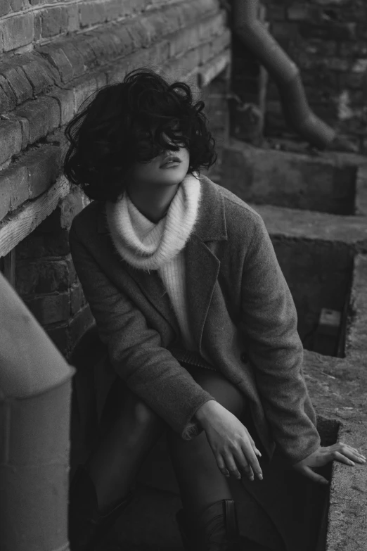 a woman in boots and a coat leaning against a brick wall