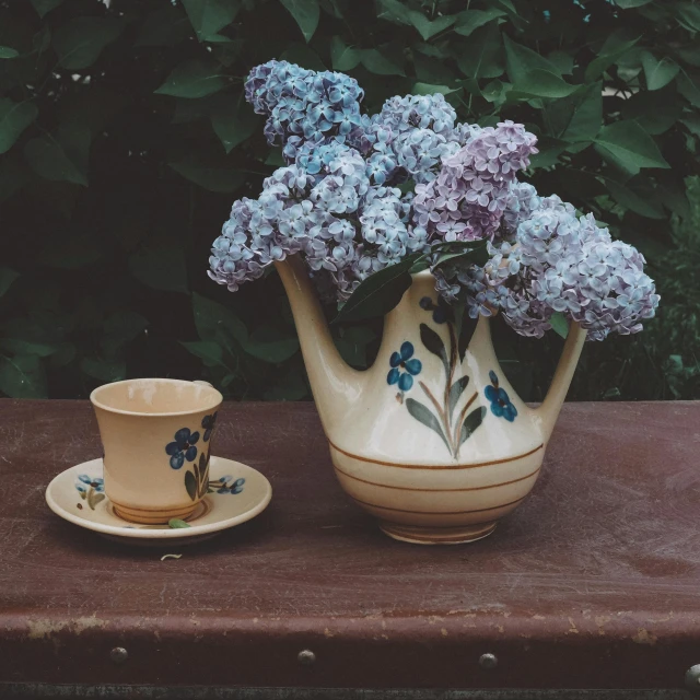 the small vase with the small blue flowers is sitting next to the large cup that has a flower in it