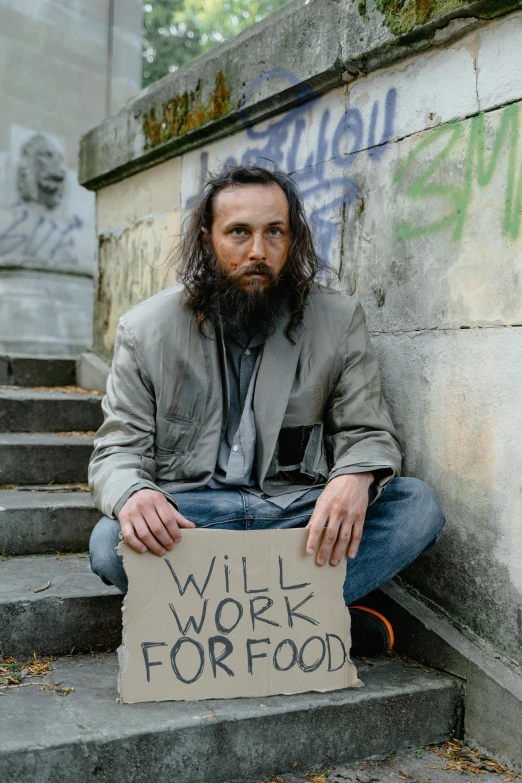 a bearded man with long hair and beard holds a sign that says will work for food