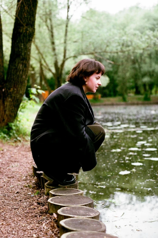 a young man kneeling on a fallen tree next to a pond