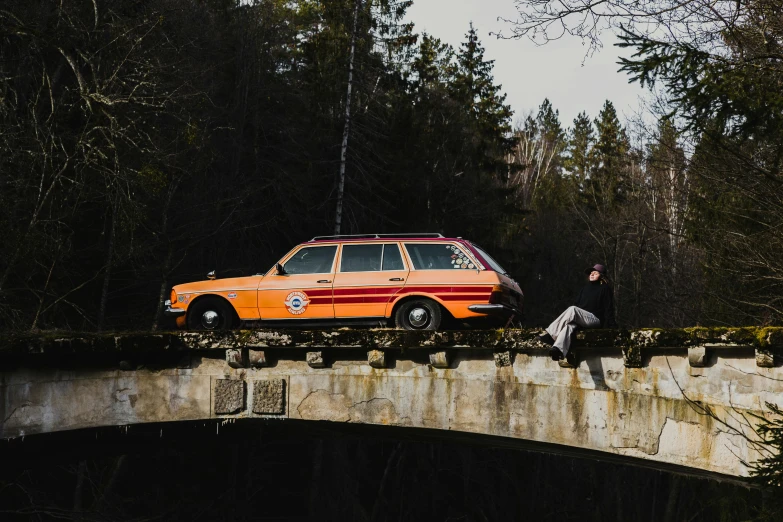 two people are sitting on the edge of a bridge with an orange and red car
