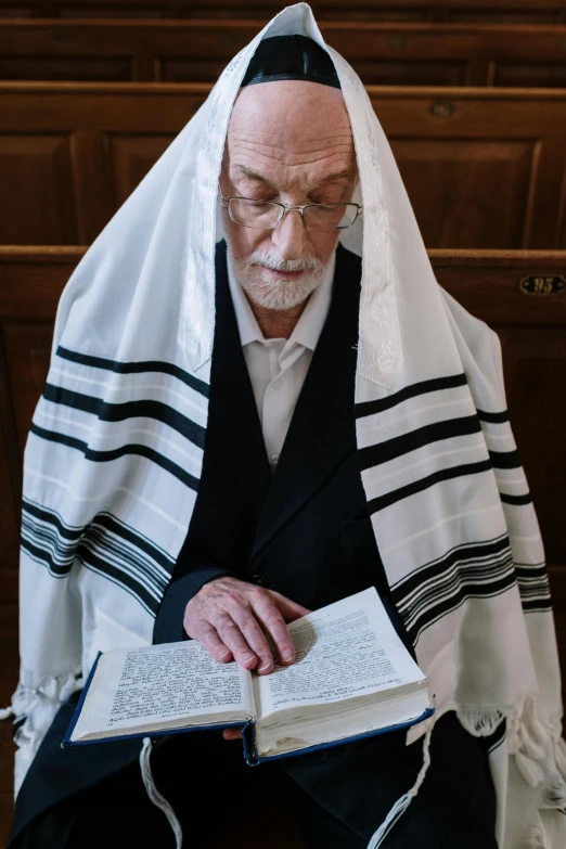 an elderly man wearing a white robe with blue accents sitting down while reading a book