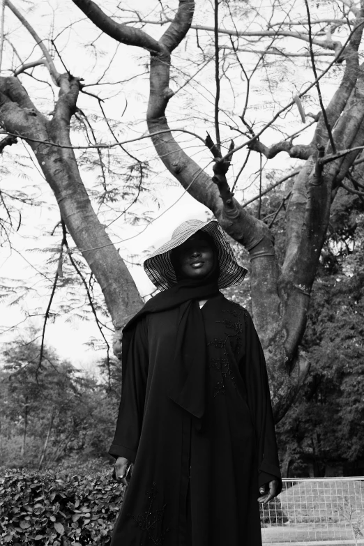 the black and white image shows a woman in a long coat with a hat and coat with beads standing in front of trees