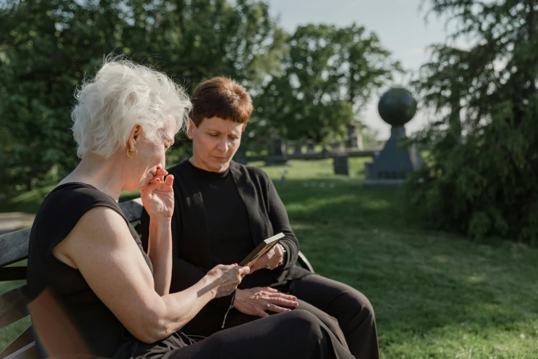 two women sitting on a bench in the park one reading a magazine and one looking at her phone