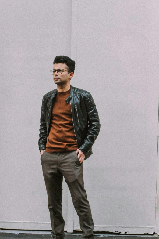 a man in glasses and a leather jacket stands against a white wall