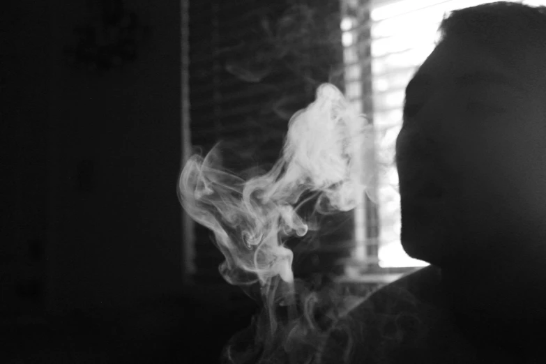 the silhouette of a man standing next to a window holding a steaming cigarette