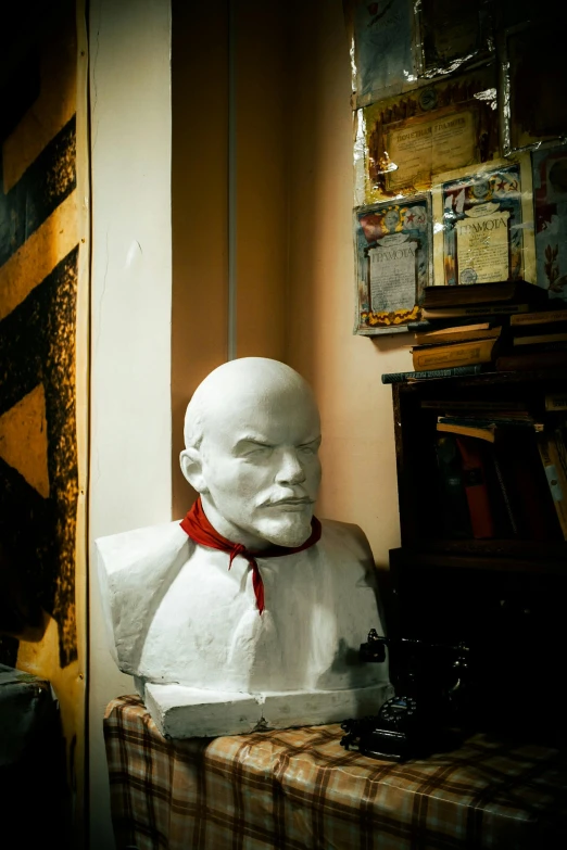 the head of a statue of a person wearing a red bow is on a wooden stand in front of a wall with books