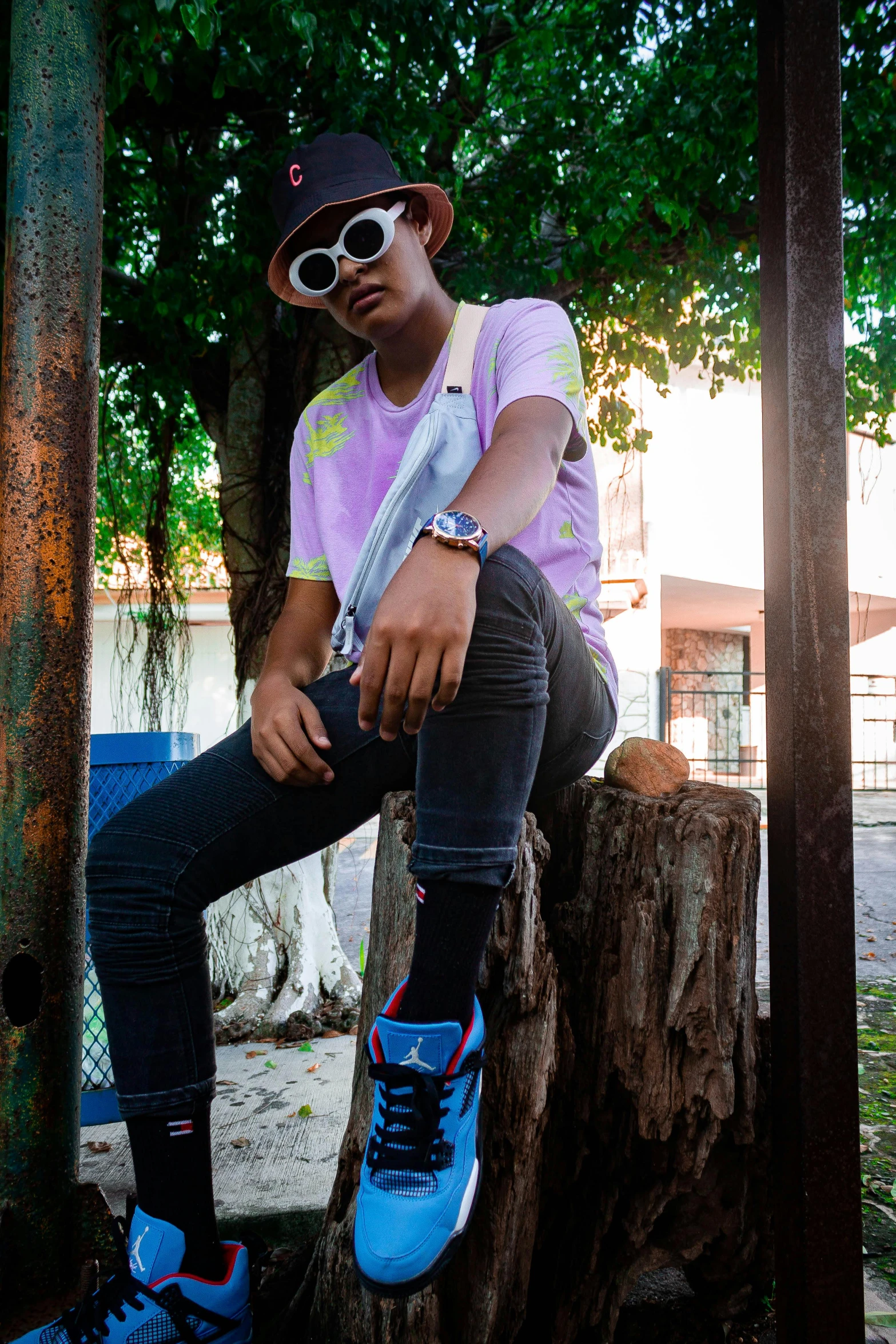 a young man wearing sunglasses and a hat sits on a tree stump