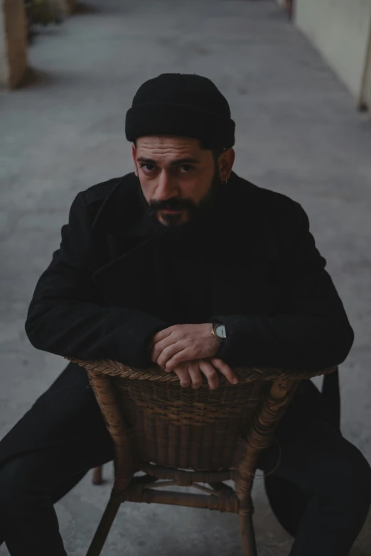 a man sitting in a wicker chair wearing a black hat and coat