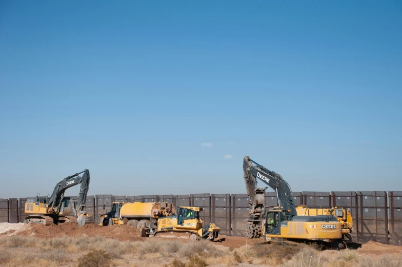 three construction equipment on a dirt surface near a fence