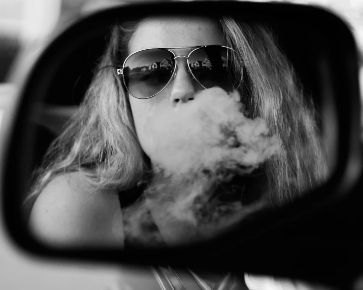 a woman with sunglasses and a cigarette is reflected in the side mirror of a car