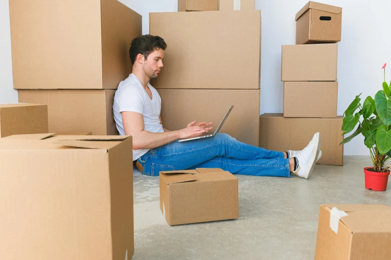 man in white shirt sitting on cardboard boxes using laptop