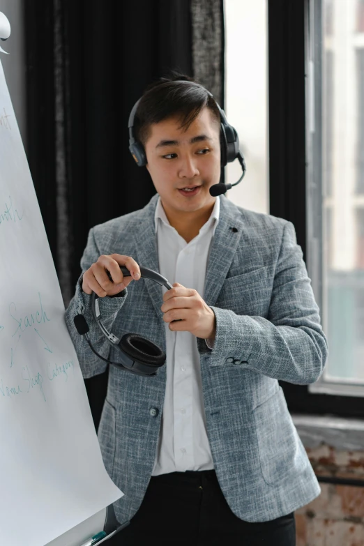 an asian male standing in front of a whiteboard with headphones on and holding a marker