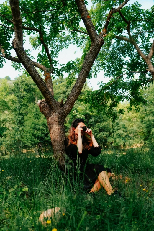 a woman sitting under a tree in a field talking on a cell phone