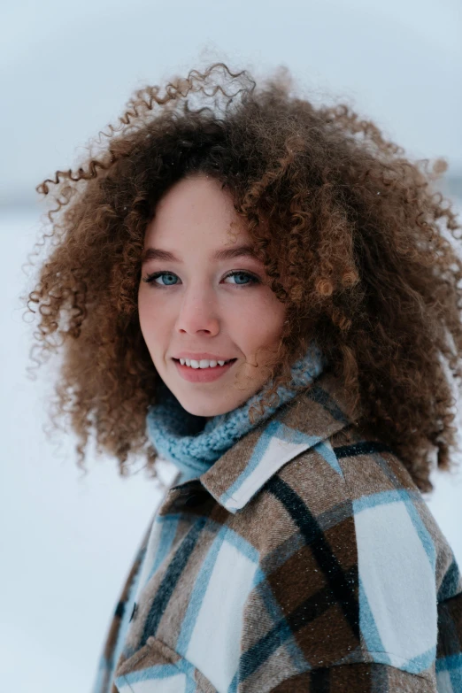 a woman with large curls and brown hair, wears a plaid coat in the snow