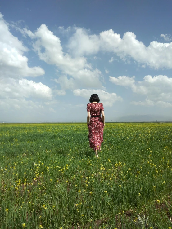 a woman walks through a wide green field with clouds overhead