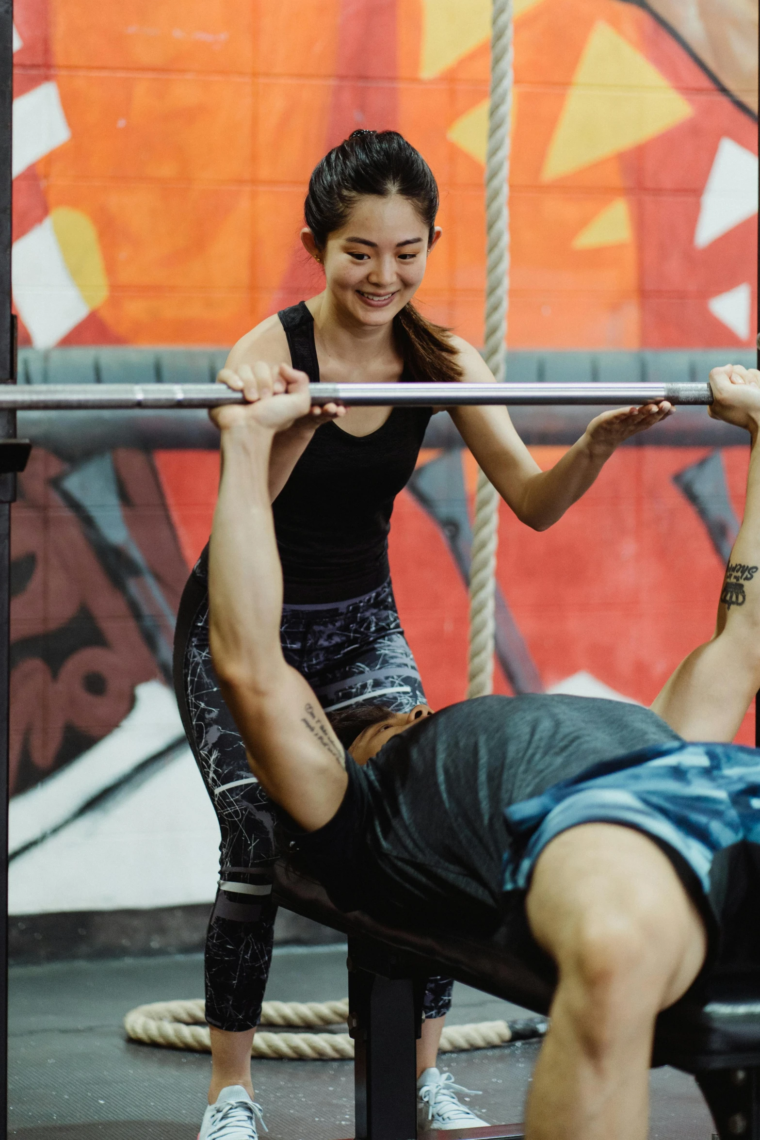 a woman lifts a bar while standing next to a man in front of a wall