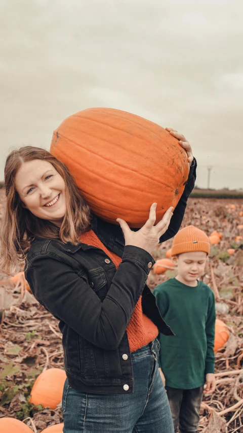 a woman holding onto an orange pumpkin with two small children behind her