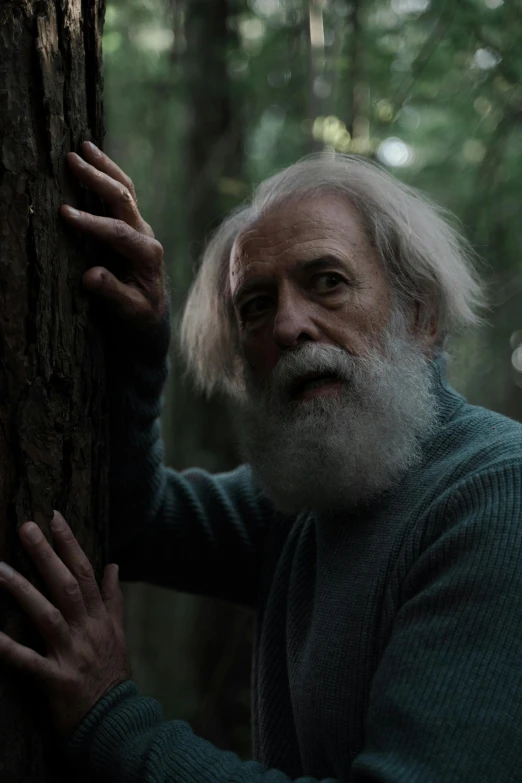 an older man with long hair, wearing a sweater leans against a tree trunk