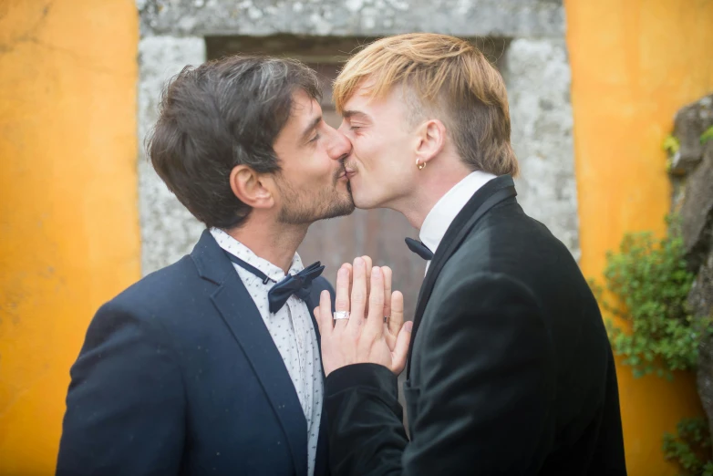 two men, dressed in suits and bow ties kiss as they stand by an orange building