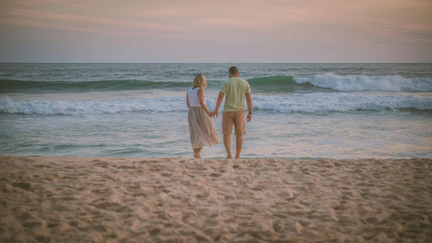 a man and woman walk towards the water holding hands