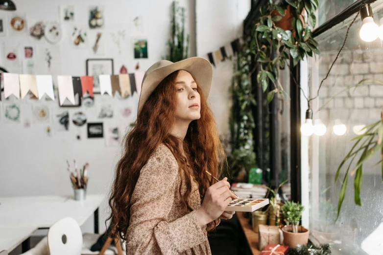 a woman with red hair in a white hat and long dress standing next to a table