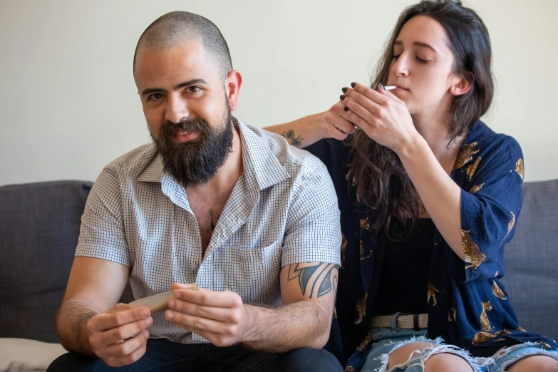 two people sitting on the couch with a woman smoking and a man brushing his beard