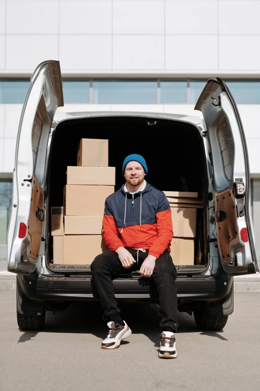 a man in blue cap and orange jacket sits inside the back of a white van filled with boxes