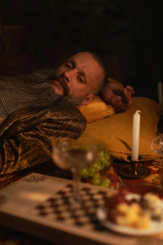 a man in medieval clothing sits on the ground next to a dinner tray with a candle