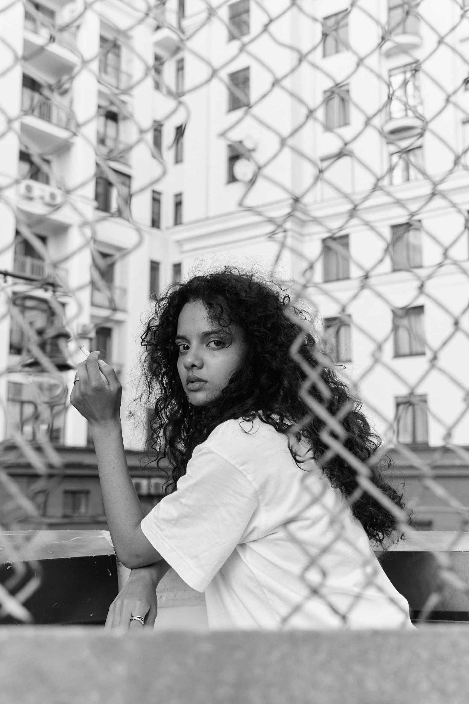 a young woman standing in front of a fence, holding a cigarette