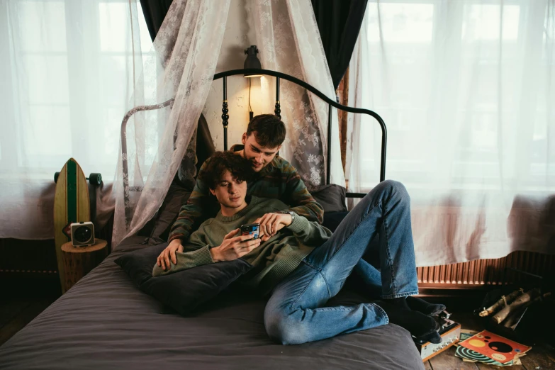 two boys are laying on the bed playing with their phones