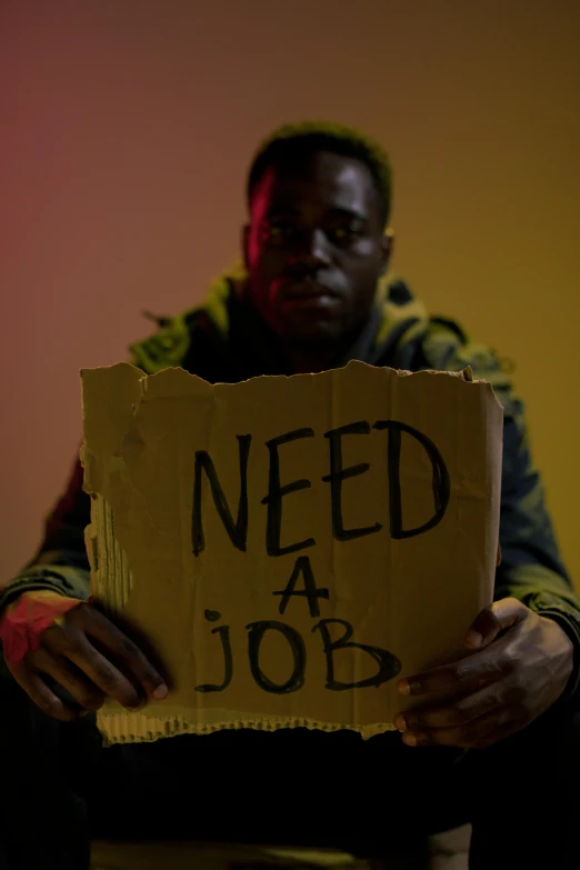 a man sitting in a room holding a sign that says need a job