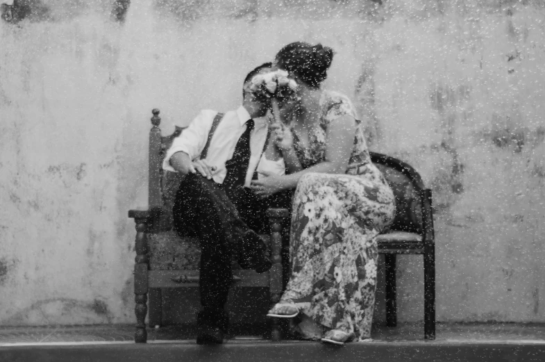 a black and white po of a woman in floral dress sitting in a chair while a man is kissing her cheek